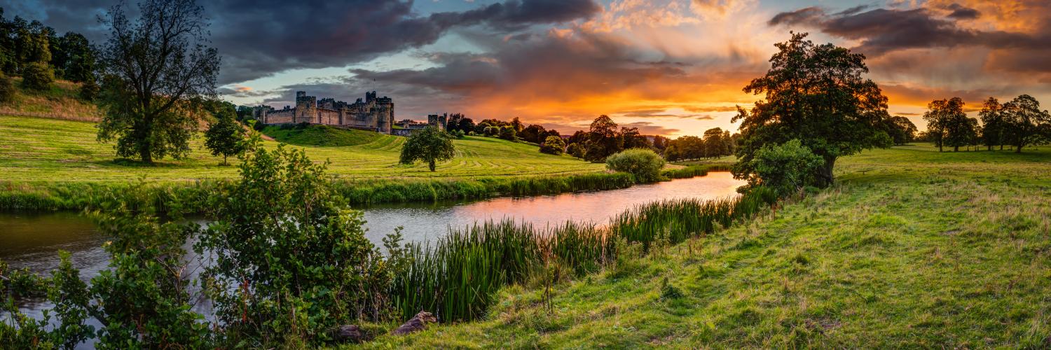 Sunset over a countryside river, surrounded by green banks with trees and a castle