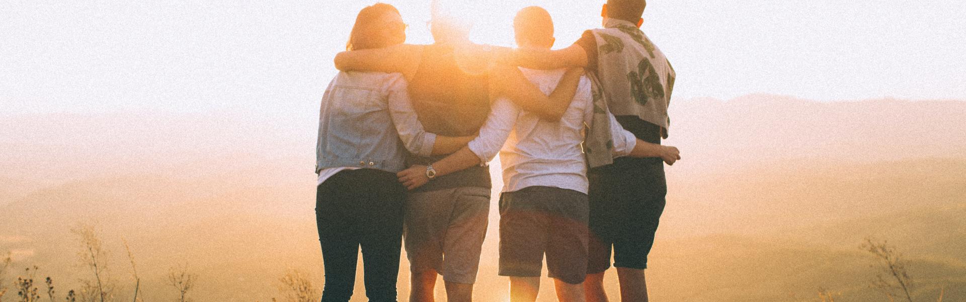 four person hands wrap around shoulders while looking at sunset