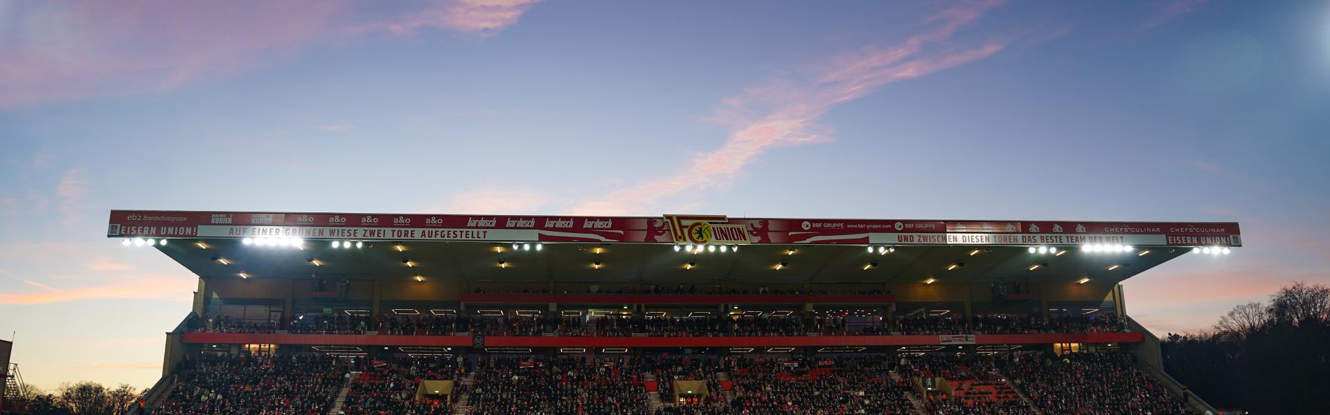 A front-on view of a football stadium and the grass pitch in front of a purple sunset
