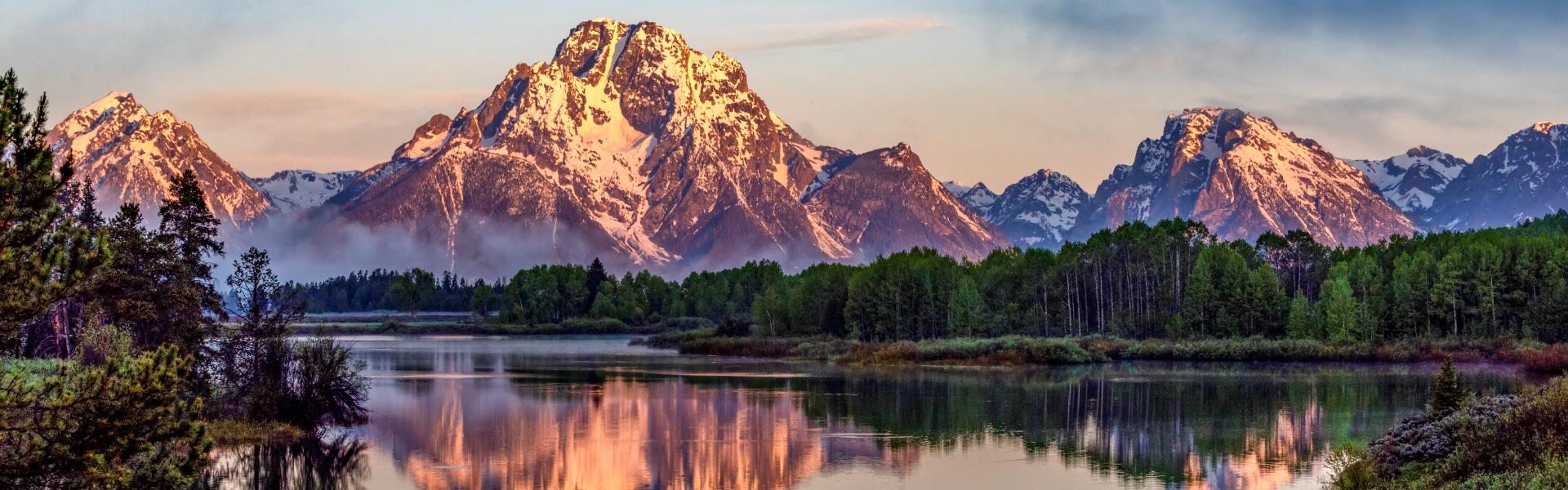 Snowy mountains forming the backdrop behind green trees and a clear lake