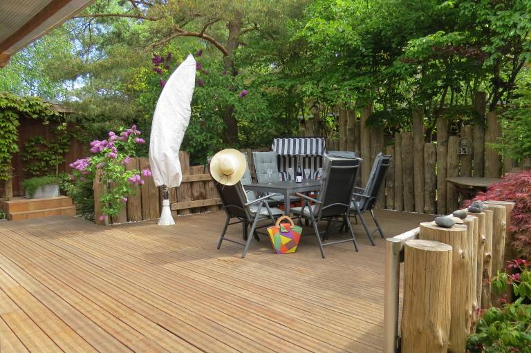 Holztisch mit Stühlen auf Terrasse, umgeben von Bäumen. [Strandhaus Möve] in [Wustrow].